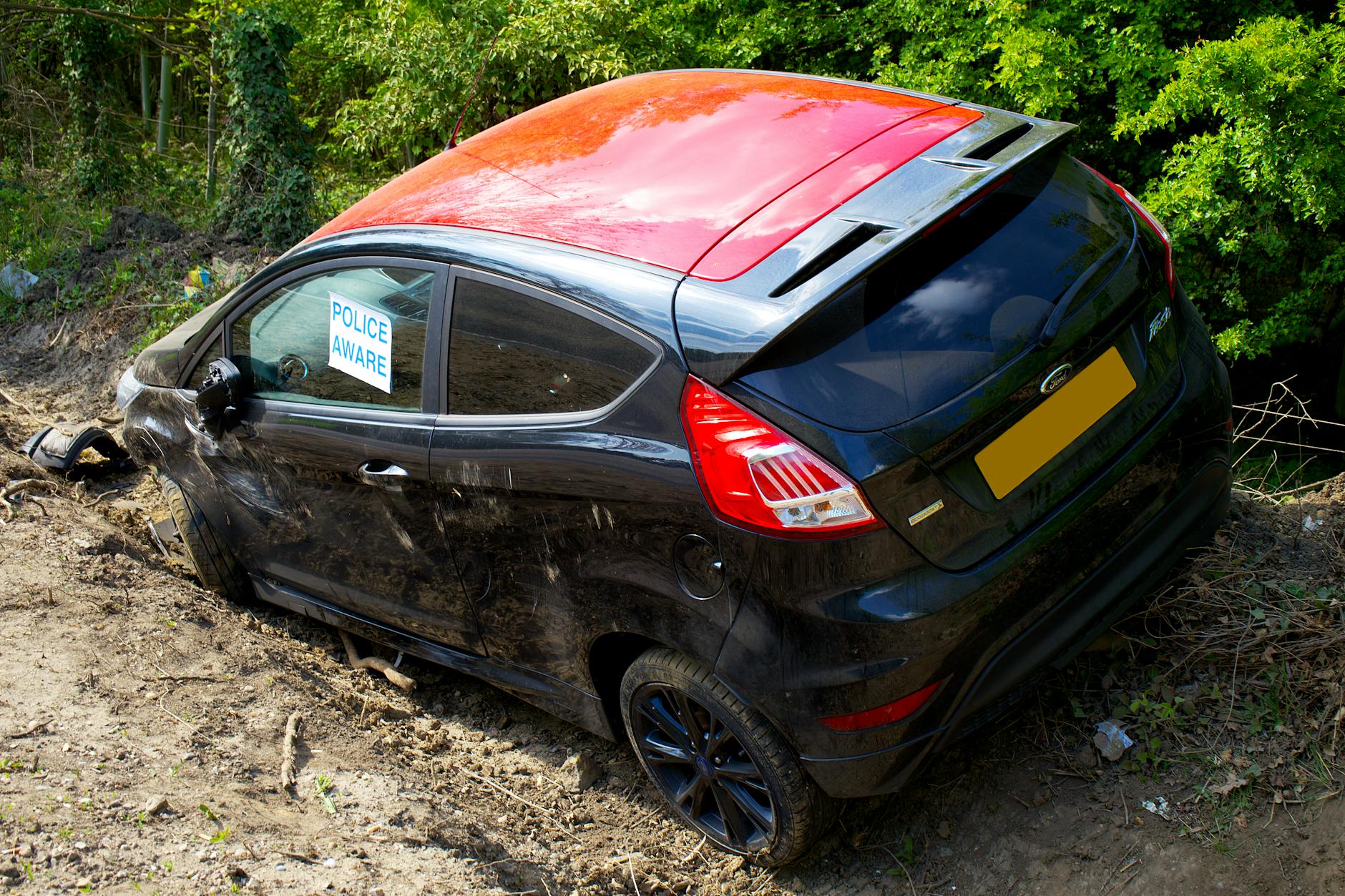 Uszkodzony czarny Ford Fiesta z tabliczką Police Aware na polnej drodze