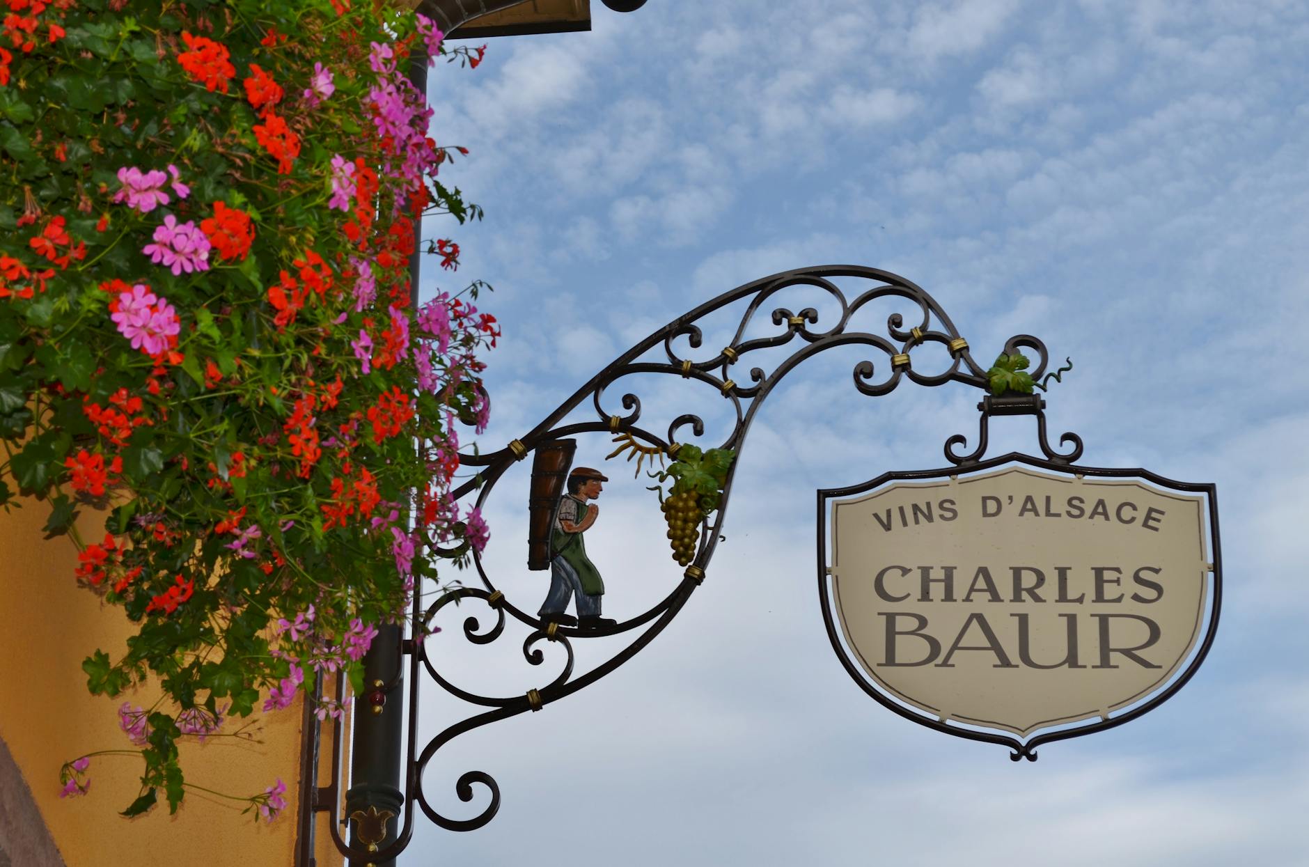 Ozdobny szyld sklepu z winem w Alzacji na tle kwiatów i błękitnego nieba